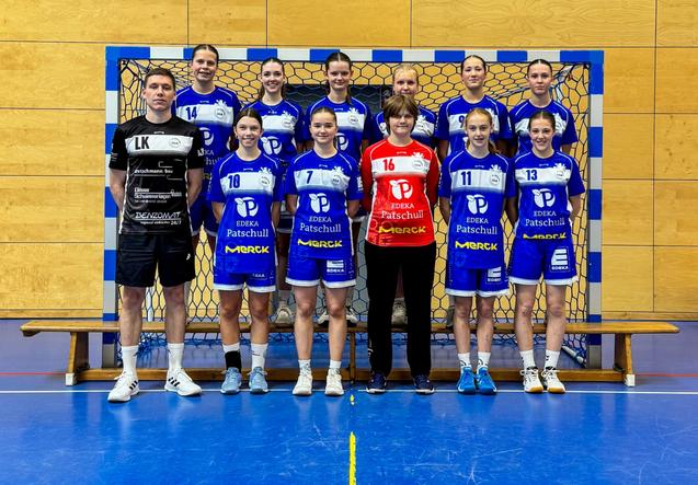 Mannschaftsbild einer Handballmannschaft in blauen Trikots vor dem Tor mit einer Trainerin und zwei Spielern in schwarzem und rotem Trikot.
