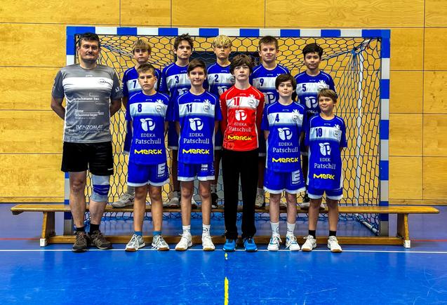 Handballmannschaft mit Trainer steht aufgestellt vor einem Tor in blauen Trikots, l&auml;chelnd und in sportlicher Pose.