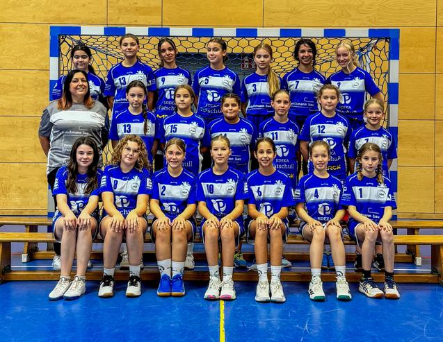 Gruppenfoto einer Handballmannschaft mit 14 Spielerinnen und Trainerin in blauen Trikots vor einem Handballtor.