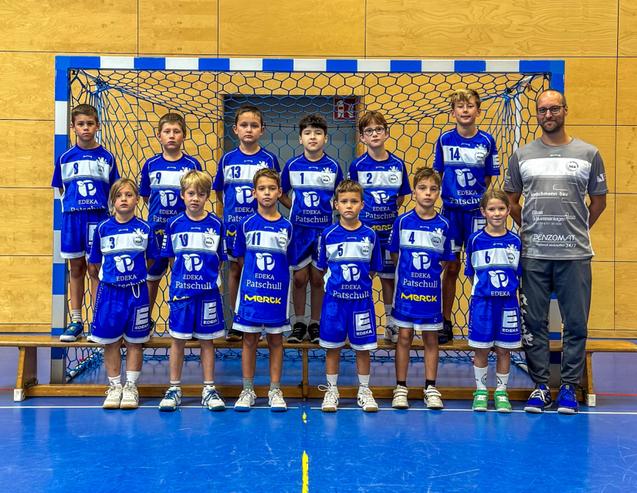 Gruppenfoto einer Handballmannschaft in blauen Trikots vor dem Tor, mit Trainer rechts im Bild.