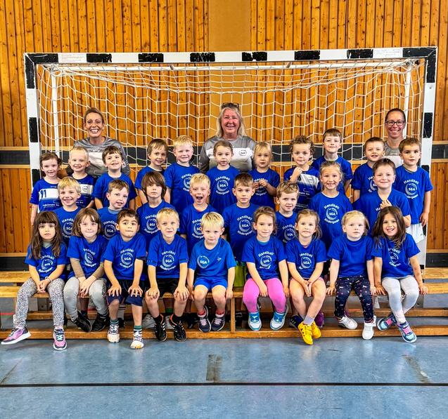 Gruppennfoto von 30 Kindern in blauen T-Shirts vor einem Handballtor, umgeben von drei Betreuern in einer Sporthalle.