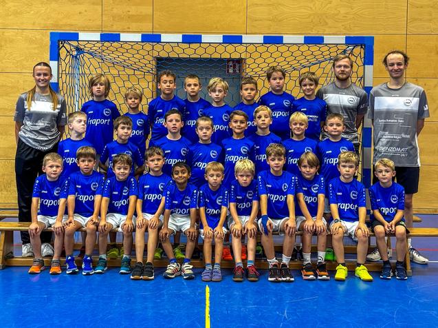 Gruppenfoto einer Handballmannschaft mit 24 Spielern in blauen Trikots und zwei Trainern vor einem Tor.