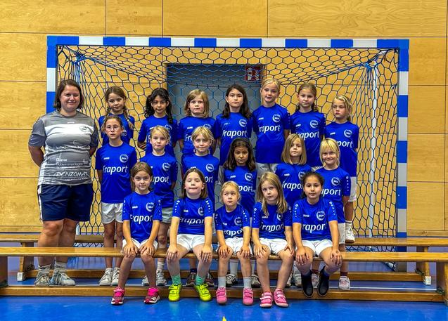 Gruppenfoto von M&auml;dchenmannschaft im Handball, Trikots in Blau mit Trainerin, vor einem Handballtor.