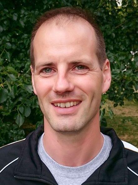 Portr&auml;t eines Mannes mit kurzem Haar, lachend vor einer gr&uuml;nen Kulisse aus B&auml;umen. Er tr&auml;gt ein schwarzes Shirt.