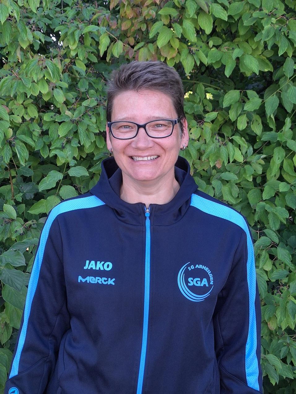 Frau mit Brille l&auml;chelt freundlich vor gr&uuml;ner Pflanzenkulisse, tr&auml;gt einen schwarzen Sportanzug mit blauen Akzenten.