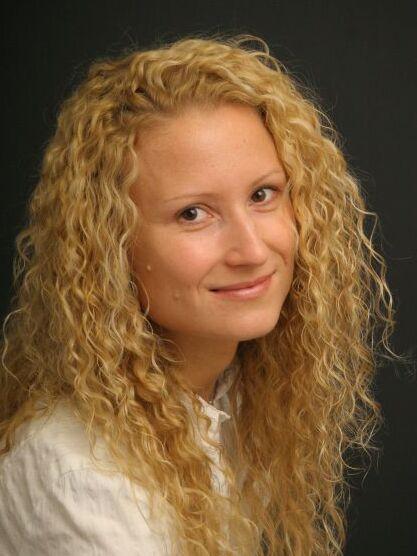 Portr&auml;t einer l&auml;chelnden Frau mit lockigem, blondem Haar vor einem dunklen Hintergrund. Sie tr&auml;gt eine wei&szlig;e Bluse.