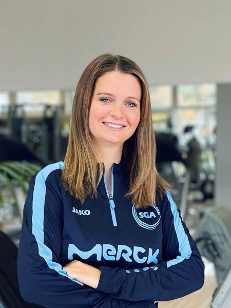 Frau mit langen Haaren in Trainingskleidung steht mit verschr&auml;nkten Armen in einem Fitnessstudio.