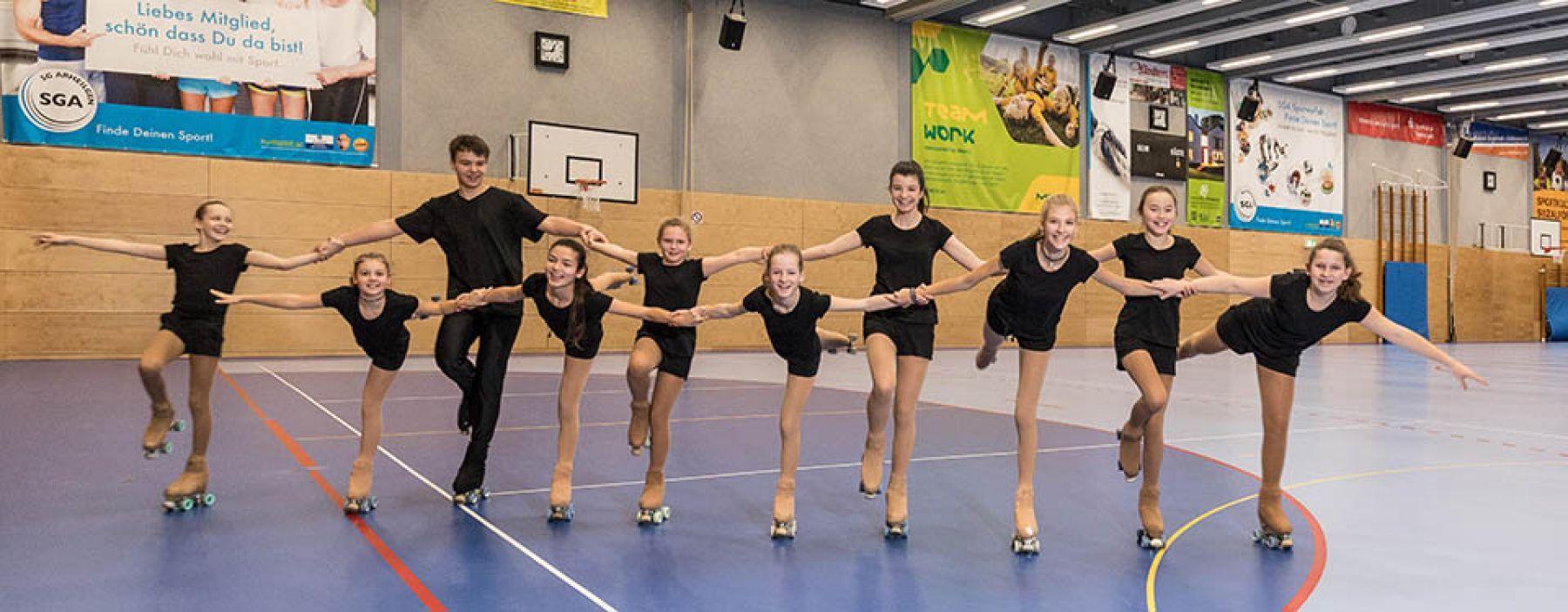 Neun Rollschuhläuferinnen in Schwarz stehen in einer Linie und halten sich an den Händen in einer Sporthalle.