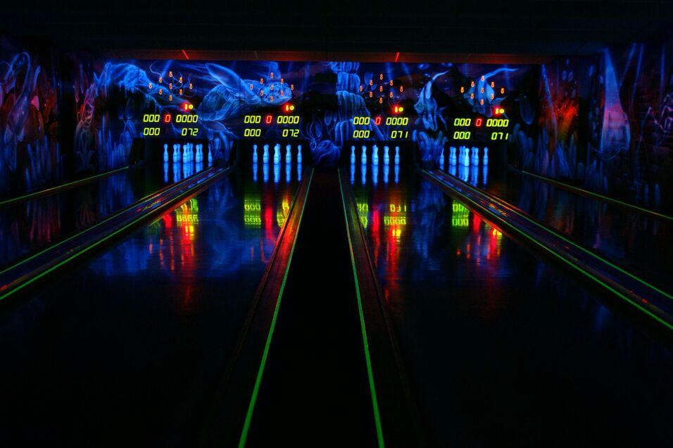 Dunkle Bowlingbahn mit buntem Neonlicht, reflektierenden Kegel und digitaler Anzeigetafel im Hintergrund.