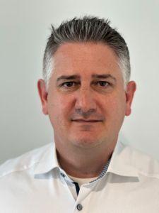 Portr&auml;t eines Mannes mit grauen Haaren, der in einem wei&szlig;en Hemd vor neutralem Hintergrund steht.