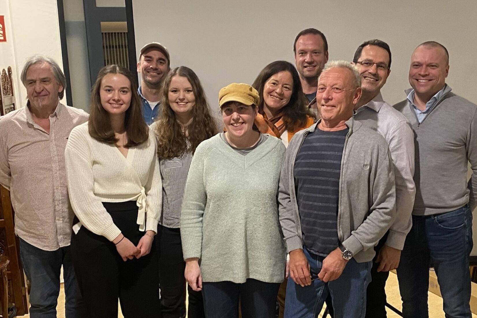 Gruppe von zehn Personen l&auml;chelnd vor einer Leinwand, stehend in einem gem&uuml;tlichen Raum mit Holzdetails.