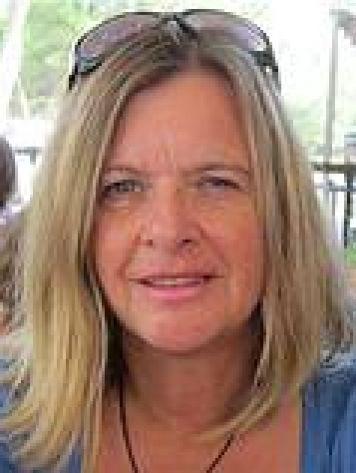 Portr&auml;t einer Frau mit langen, blonden Haaren und einer Sonnenbrille auf dem Kopf, l&auml;chelnd vor unscharfem Hintergrund.