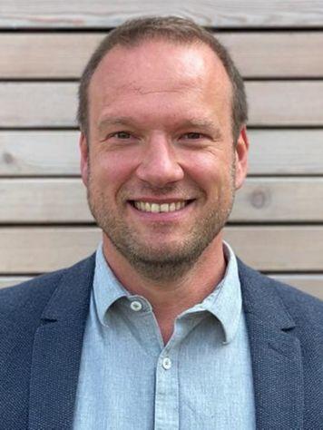 Nahaufnahme eines l&auml;chelnden Mannes in einem blauen Blazer vor einer Holzlattenwand.