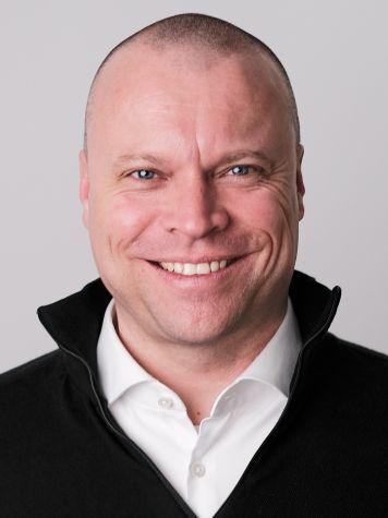 Portr&auml;t eines l&auml;chelnden Mannes mit Glatze, in einem schwarzen Pullover und wei&szlig;em Hemd. Hintergrund in hellem Grau.