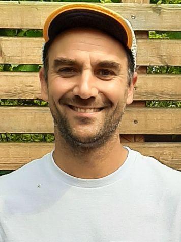 L&auml;chelnder Mann mit kurzen Haaren und M&uuml;tze, vor einer Holzlattenwand. Tr&auml;gt ein einfaches, helles T-Shirt.