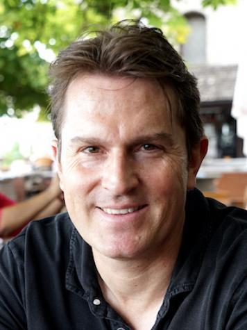 Portrait eines l&auml;chelnden Mannes mit kurzen, braunen Haaren, sitzend in einem Caf&eacute; unter gr&uuml;nen B&auml;umen.