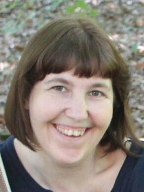 L&auml;chelnde Frau mit schulterlangen, braunen Haaren, vor unscharfem, bl&auml;tterigem Hintergrund.