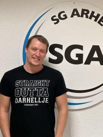 L&auml;chelnder Mann in schwarzem T-Shirt mit Aufdruck vor dem Logo der SG Arheilgen an einer wei&szlig;en Wand.