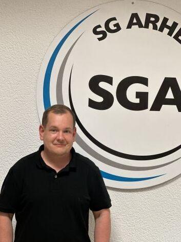 Mann in schwarzem Shirt steht vor einem runden Vereinslogo der SG Arheilgen an einer Wand.