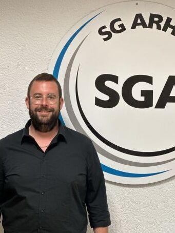 Mann in schwarzem Hemd steht neben einem runden Logo der SG Arheilgen mit der Aufschrift "SGA".