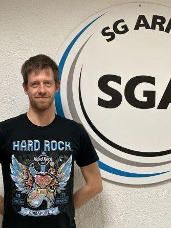 Mann mit T-Shirt steht neben einem runden Schild mit dem Text &bdquo;SG Arheilgen SGA&ldquo; an einer Wand.