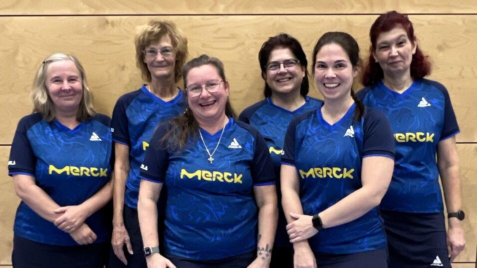 Sechs Frauen in blauen Sporttrikots mit gelbem "Merck"-Logo, l&auml;chelnd vor einer Holzwand.