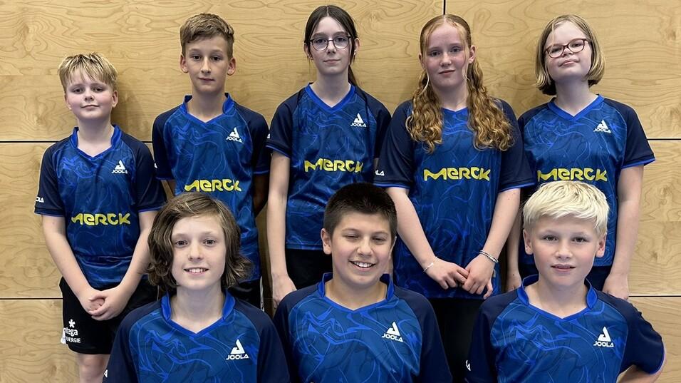Gruppenshooting von acht Kindern in blauen Tischtennistrikots mit &bdquo;Merck&ldquo;-Aufdruck vor einer Holzwand.