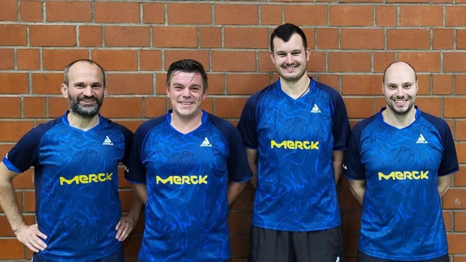 Vier Tischtennisspieler in blauen Trikots mit Merck-Logo stehen vor einer Ziegelwand und l&auml;cheln in die Kamera.