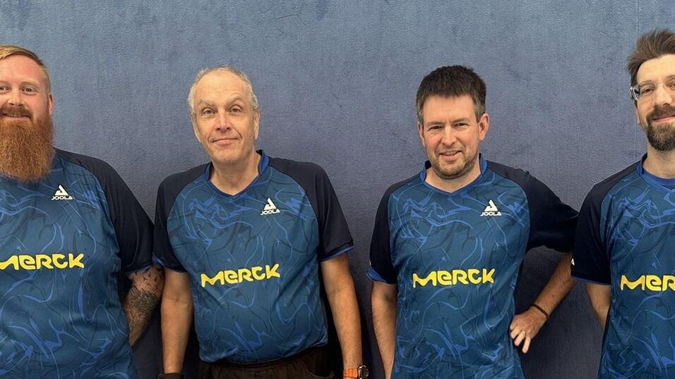 Vier M&auml;nner in blauen Tischtennistrikots stehen nebeneinander vor einer grauen Wand.