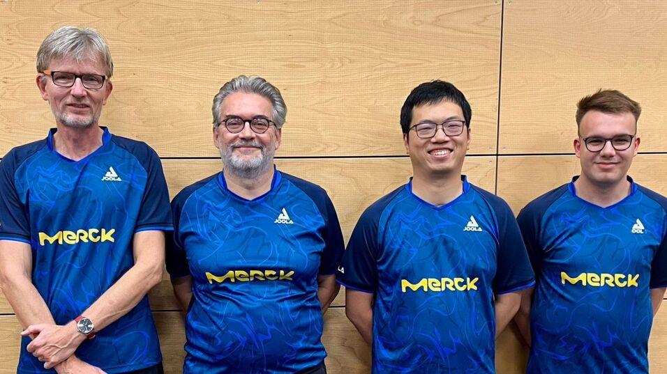 Gruppenfoto von vier Personen in blauen Sporttrikots mit "Merck"-Aufdruck vor einer Holzwand.