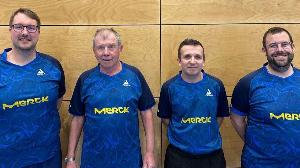 Vier m&auml;nnliche Tischtennisspieler in blauen Trikots mit gelbem Aufdruck "Merck" stehen vor einer Holzwand.