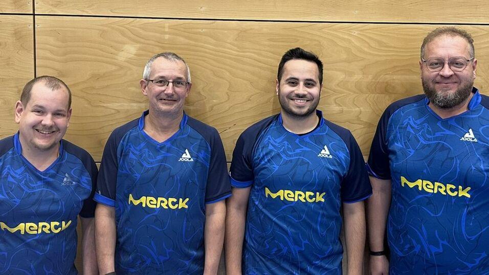 Vier Tischtennisspieler in blauen Trikots mit dem Logo &bdquo;Merck&ldquo; stehen l&auml;chelnd vor einer Holzwand.