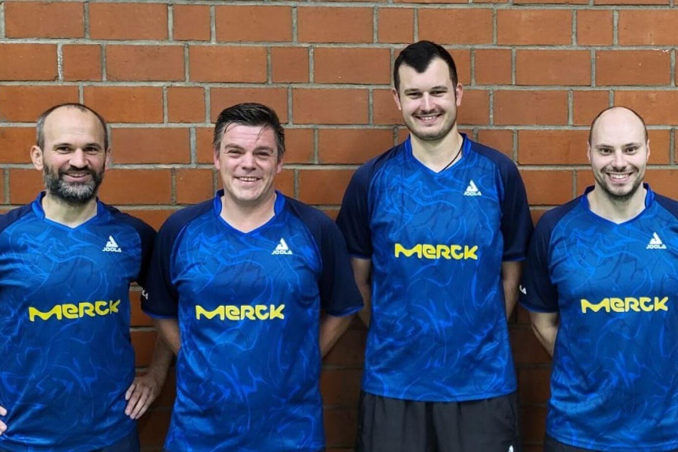 Vier Tischtennisspieler in blauen Trikots mit Merck-Logo stehen vor einer Ziegelwand und l&auml;cheln in die Kamera.