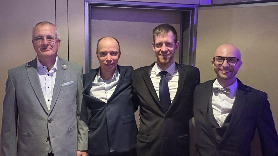 Vier M&auml;nner in Anz&uuml;gen stehen l&auml;chelnd in einem festlichen Raum vor einer Wand mit sanftem Licht.