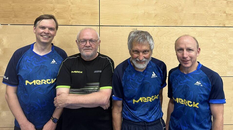 Vier Tischtennisspieler posieren l&auml;chelnd in blauen Trikots mit der Aufschrift &bdquo;Merck&ldquo; vor einer Holzwand.