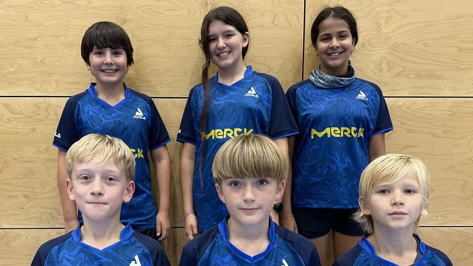 Sechs Kinder in blauen Tischtennis-Trikots mit gelbem "Merck"-Logo stehen vor einer Holzwand.