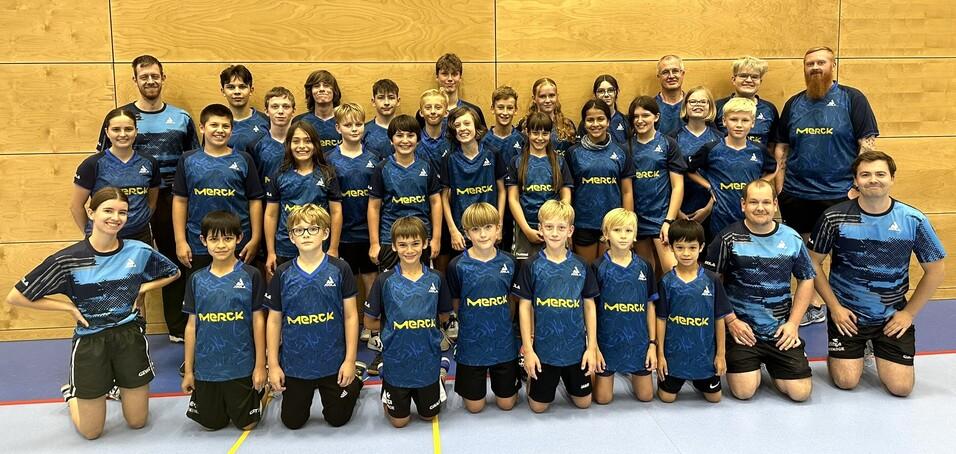 Gruppenfoto von Jugendlichen und Erwachsenen in blauen Trikots mit "Merck"-Aufdruck in einer Sporthalle.