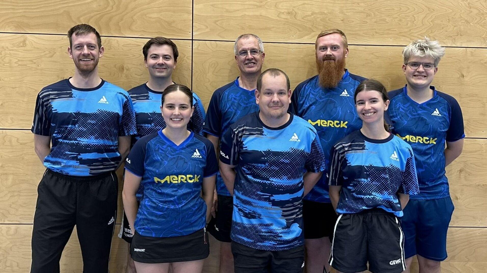 Gruppe von acht Tischtennisspielern in blauen Trikots vor einer Holzwand, l&auml;chelnd f&uuml;r das Foto aufgestellt.