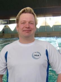 Mann mit blonden Haaren in einem wei&szlig;en Shirt steht vor einem Schwimmbecken. Hintergrund zeigt Schwimmbahnmarkierungen.