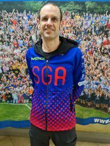 Mann in blauer Jacke mit rotem Schriftzug &bdquo;SGA&ldquo; vor einer Menschenmenge auf einem Banner.