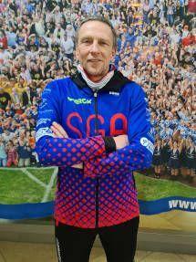 Mann mit verschr&auml;nkten Armen in einer bunten Sportjacke vor einem Hintergrund aus jubelnden Fans.