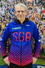 Mann in blauer, rot gemusterter Trainingsjacke mit "SGA"-Aufdruck steht vor einer Menschenmenge.