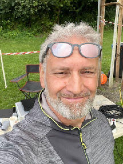 Mann mit grauen Haaren und Bart l&auml;chelt in die Kamera, im Hintergrund ein gr&uuml;ner Rasen und einige Gegenst&auml;nde.