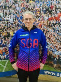 Mann in blauer, rot gemusterter Trainingsjacke mit "SGA"-Aufdruck steht vor einer Menschenmenge.