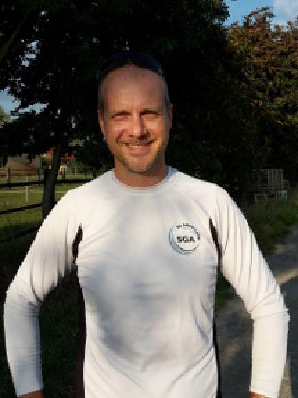 L&auml;chelnder Mann in einem Langarmshirt steht auf einem Weg, umgeben von B&auml;umen und Vegetation.