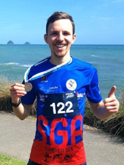 L&auml;ufer in blauem Shirt zeigt stolz eine Medaille an einem K&uuml;stenweg mit Meer im Hintergrund.