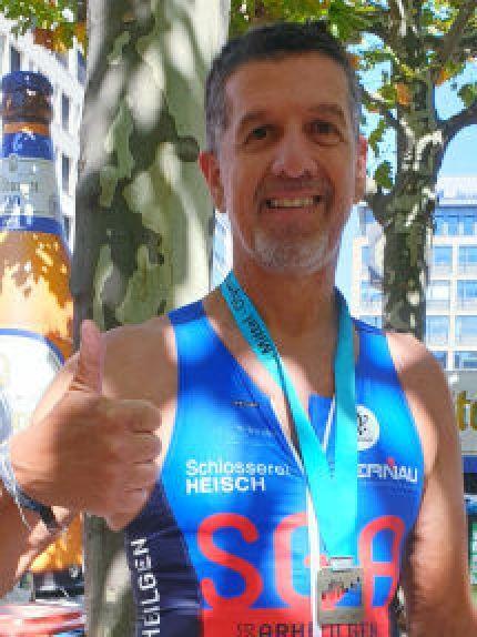 Mann in Sportkleidung zeigt Daumen hoch, tr&auml;gt Medaille, hinter ihm eine stark stilisierte Bierflasche und B&auml;ume.