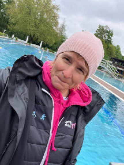 Frau mit M&uuml;tze und Jacke steht am Beckenrand eines Schwimmbads mit Wasser und B&auml;umen im Hintergrund.