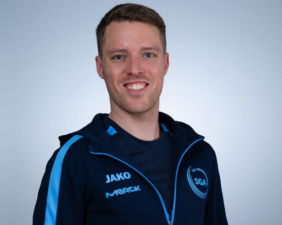 Mann mit kurzen Haaren, Trikot der SGA Arnecken mit blauem Logo, steht vor gr&uuml;nem Hintergrund aus Pflanzen.