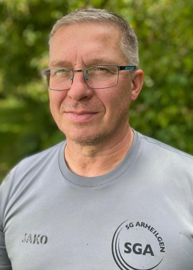 Portrait eines lächelnden Mannes mit Brille, in grauem Shirt mit Aufdruck, vor einer grünen Pflanzenkulisse.